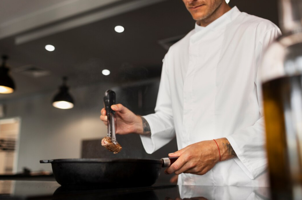 Chef in white coat cooking with tongs over a hot pan in a kitchen setting